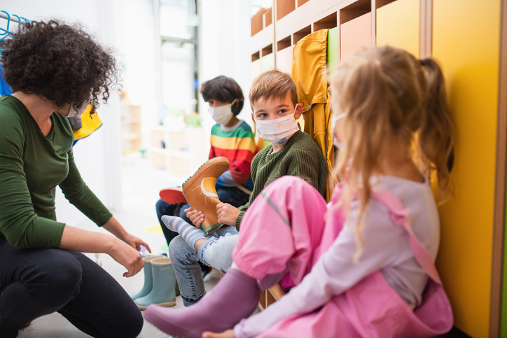 Pre school teacher helping children to put on shoes indoors in cloakroom at nursery, coronavirus concept.