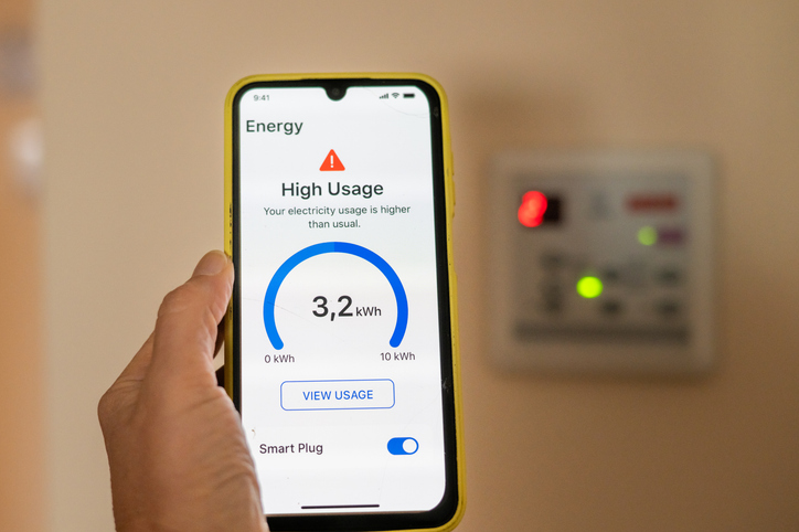 Hand holding smartphone displaying high electricity usage alert from smart plug application, with blurred electricity meter in background, promoting energy saving and cost reduction