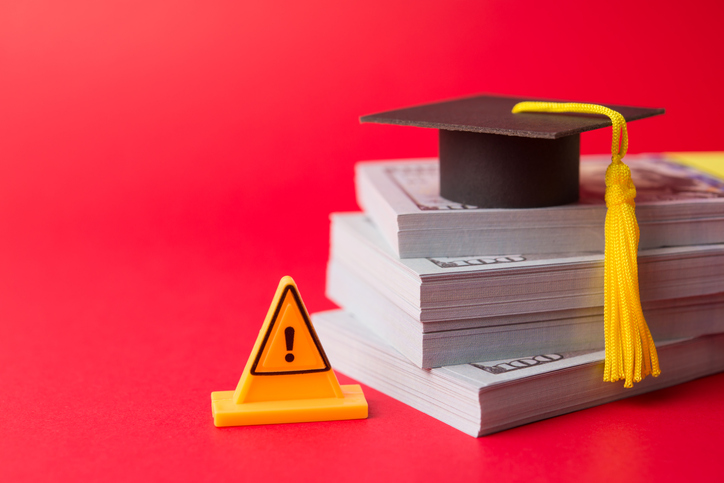 Yellow warning sign, graduation hat mortarboard, heap US dollar banknotes on red background.